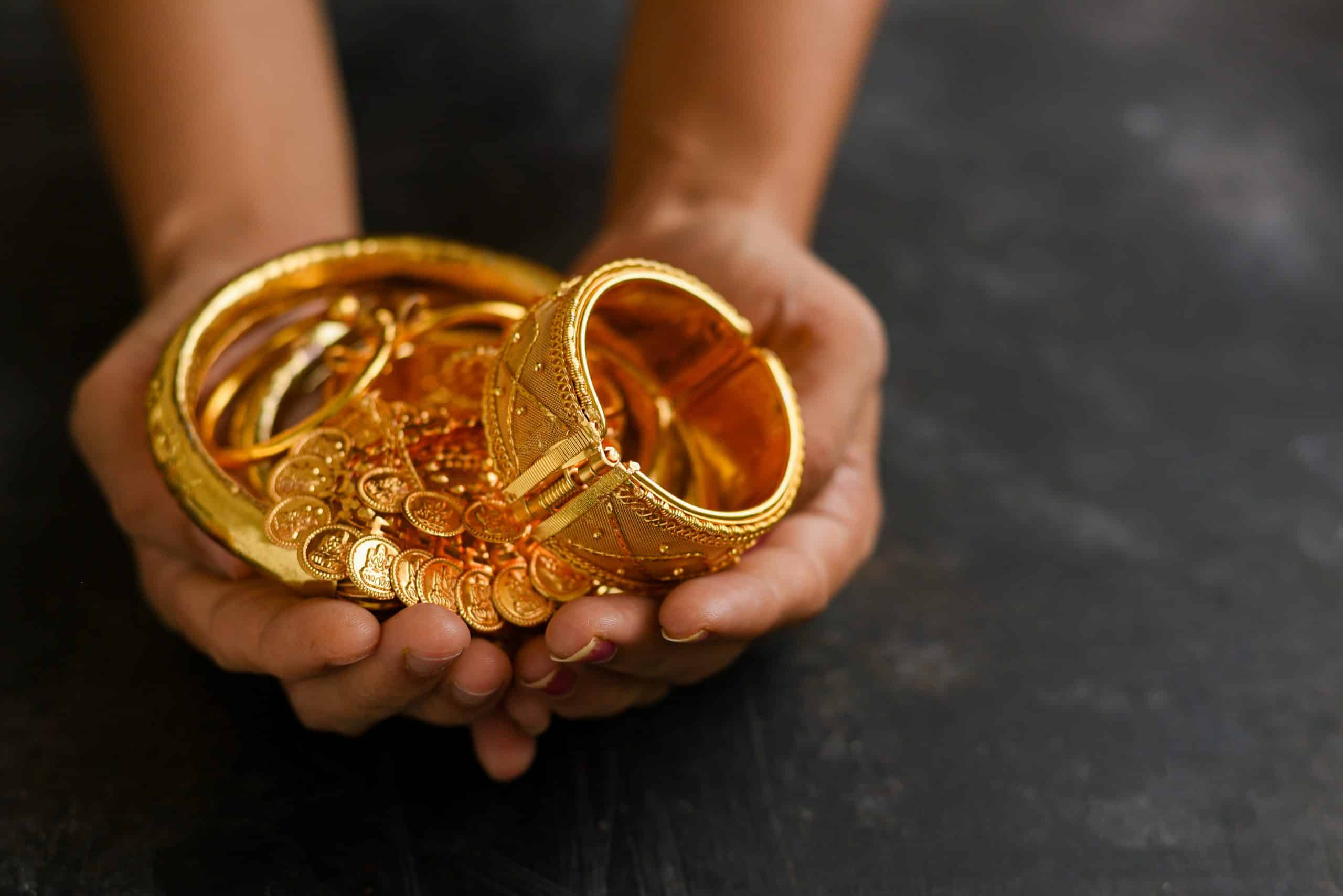 Hands holding a collection of traditional yellow gold jewelry, including bangles, bracelets, and gold coin ornaments, displayed over a dark background.